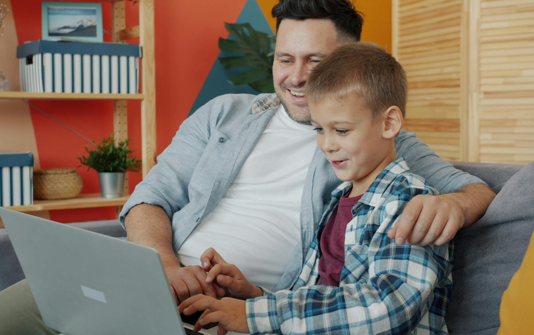 Father and son using laptop computer talking sitting on couch at home together surfing the internet.